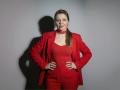 VeronikaGoldy with hands on hips, wearing a red suit, red lace choker, and red top, shadow on gray wall, brown ponytail, red lipstick, confident smirk.