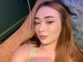 JaneKrover has light brown hair, winged eyeliner, and pink lipstick. She's wearing a beige top, lying on a pinkish-orange surface, with a subtle necklace. Her skin is shiny.