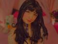 AnnieGrey with black wavy hair, plaid choker, and tattoos, smiling at the camera. She has red lipstick and eyeliner. Boobs visible, pink bed in the background.