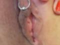 La vulve de Shanna, légèrement écartée par les doigts, révèle ses petites lèvres roses, humides et légèrement ridées, avec une bague en argent à un doigt. On peut voir son ongle peint en rouge..
