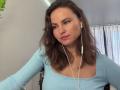 TangelaEmmel with wavy brown hair, light blue ribbed shirt, white earbuds, pink lipstick, and a slight pout, looking directly at the camera. Boobs visible under the shirt.