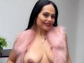 CharlotteCrofort with long black hair, red lipstick, and pink furry vest, exposing her big, bare, brown-tipped tits, smiling, wearing hoop earrings, and a pink necklace.