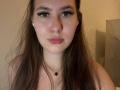 DeborahHaughey has long brown hair, green eyes with thick eyelashes, pink lips, fair skin, small black cross necklace, and white earbuds, looking straight at the camera.