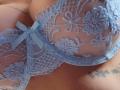 FrenchShanna's tits in a sheer, blue lace bra with floral patterns and a small blue bow. Her tan skin and tattoo are visible. Close-up, lingerie, sexy.