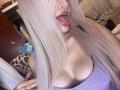 EliseMonet with long pinkish-blond hair, tongue out, purple tank top, showing big tits, pale skin, in a bedroom. Her hand touches her hair, she's looking at the camera.