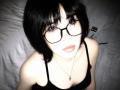 ChantellHoulihan with black short hair, glasses, and a black tank top, looking up with pale skin, full lips, and a necklace, sitting on a bed.