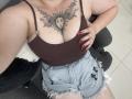 DiLaria with large boobs, brown tank top, and frayed denim shorts, showing a sun face tattoo between her tits, red nails, sitting on a gray chair.