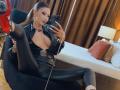 LucyGrace with her tits barely covered in a black leather outfit, holding her phone, legs spread, showing her red high heel and a long chain necklace, sitting on a chair.