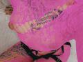 DenyChantal wearing a pink lace top with large cutouts, revealing her tits, and black strappy bottoms, showcasing a tattoo on her right thigh. Pink lace covers her pussy.