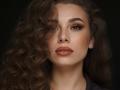 Niferttiti has curly brown hair covering one eye, full lips with pinkish-brown lipstick, and smoky eye makeup. Her expression is serious with a slight, seductive, confident look.