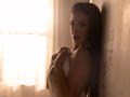 KarlaSoto with wet brown hair, holding a white towel against her tits, standing in a sunlit, beige-walled room, looking over her shoulder with a sultry expression.