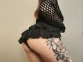 Niferttiti wearing a black fishnet top and ruffled black skirt, exposing her bare ass and a large floral tattoo on her right thigh.