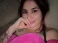 AnaEox with dark hair, a nose ring, and a lip piercing, holding a pink blanket, showing tattoos on her chest and arm, with a subtle smile.