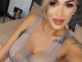 NicolSNaya has blonde hair, thick eyebrows, and red lipstick. She's wearing a sheer blue top, showing her big tits, and has tattoos on her arms. She's smiling at the camera.
