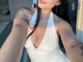 Olieri with long black hair, wearing a white halter dress, showing her tits in a sunlit room, smiling, has a tattoo on her right arm.