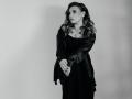 Niferttiti with wavy hair, wearing a black dress and leather jacket, standing with hands crossed, looking to the side, in a black and white photo. Boobs visible under dress.