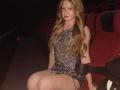 AkilahGhil with long blonde hair, wearing a sparkly, one-shoulder black dress, sitting in a red theater seat, showing her bare thighs and subtle tattoo on her right leg.