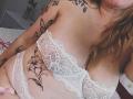 PauleetteRoux has large tits, wearing a white lace bra and panties, with butterfly and lily tattoos on her chest and belly, her blonde hair partially visible.