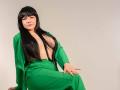 AmarantaSoto with long black hair, green outfit, and exposed tits, sitting with one hand on her green skirt, looking at the camera. Soft lighting.