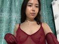 MiiaVixen with medium tits, wearing a sheer red top, showing nipples, no bra, long dark hair, light skin, pouty lips, necklace, standing in front of a green curtain.