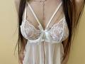 Tisia's white lace babydoll with strappy details and a ribbon, showing her tits through sheer fabric, long brown hair, and a delicate necklace against a yellow background.