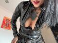 LiaMiss draagt een glanzende zwarte latex outfit, waarvan de rits gedeeltelijk open is, waardoor haar grote borsten en getatoeëerde borst zichtbaar zijn. Ze heeft rode lippenstift en een neusring, en zwart haar..