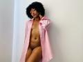 RoldanStefy with curly black hair, wearing an open pink shirt, exposing her small tits, dark brown pussy, and bare brown skin, standing against a white wall.
