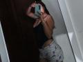 AntoniaBrunni with wet brown hair, wearing a black tank top and white pajama shorts with purple patterns, taking a mirror selfie, showing her ass and small tits.