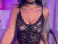 MajoJones wearing a black lace lingerie top with deep cleavage, showing tattoos on her chest and arms, and a black choker with a ring. Her tits are visible.