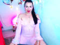 EmelySofia has pale pink off-shoulder dress, showing small tits, sitting with legs apart, red lipstick, black hair in ponytail, blue and pink light, heart necklace, smiling.