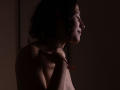 MaryCruzz with curly black hair, smiling side profile, topless, holding red flower, dark shadowed room, soft light, visible nipples, slight shadow on her tits, subtle smile.
