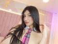 NaiaSanders with long black hair, nose ring, pink and black plaid tie, fishnet top, and exposed tits, smiling at the camera, holding her hair.