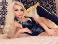 Mademoiselle Bella, com cabelo loiro platinado, um arnês de couro preto e botas pretas com cadarço, deitada em uma cama rosa, mostrando seus seios e nádegas, usando um colar de ouro..