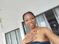 LilNaughtySally with dark skin, braided hair, gold earrings, and a blue towel covering her tits, smiling, hand with red nails on chest, standing in front of glass doors.