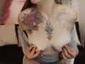 ArianaBlonde with tattoos on her chest, holding her tits with red-nailed fingers, wearing a gray top, exposing a rose, floral, and central black design on her breasts.