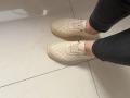 Elliance's feet in beige quilted sneakers, wearing dark gray jeans, standing on white tiled floor. Visible knees and lower legs.