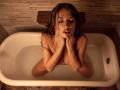 KarlaSoto, naked, with wet brown hair, sits in a white bathtub, hands on her chin, eyes closed, water covering her tits and pussy, in a warm, brown-tiled bathroom.