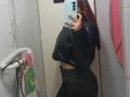 AntoniaBrunni in a black leather jacket and black pants, showing a side view of her ass in a mirror selfie, holding a blue phone, with brown hair.