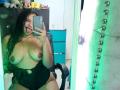 AriaVega with curly black hair, tongue out, holding phone, topless, showing large bare boobs, black shorts, sitting in chair, mirror selfie, green light reflection.