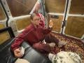 HotCaramelsTrio with glasses, in a red track jacket and maroon pants, sitting on a black leather couch, holding a beer bottle and a white mug, in a van.