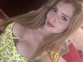 EliseMonet with long blonde hair, wearing a yellow floral dress, showing her large tits, red lipstick, and eyelash extensions, smiling at the camera.