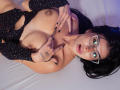 AmilyBella with glasses, black polka-dot top, and black nails, lying on a bed, exposing her titties, with tattoos on her fingers, and a surprised expression.