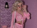 MademoiselleBella with blonde curls, pink eyes, and a sexy smile, wearing a sheer pink top, showing her tits, holding pink sunglasses, pink wall, and pink patterned background.