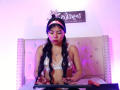 CataMoreno with long black hair, wearing a white bra and headband, sitting on a bed with a tufted headboard, typing on a keyboard, her tits visible, a dildo on the bed.