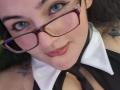 LindaSophieX with glasses, blue eyes, and red lipstick, wearing a black and white outfit with a collar, showing her tits slightly, smiling at the camera.