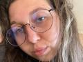 Sanella with puffy blue-tinted glasses, messy brown hair, and a slight pout, showing a small zit on her nose, looking directly at the camera.
