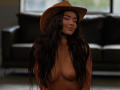 EmmaNoor with long wavy brown hair, brown cowboy hat, and pink eye makeup, topless, showing her small, perky tits and a slight smile, standing in a dim room.