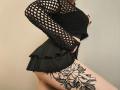 Niferttiti in a black fishnet top and flirty skirt, showing a large floral tattoo on her right thigh, with her ass and tits visible in profile.