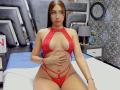 EmmaRubis with long brown hair, wearing a red lace halter top and matching strappy panties, covering her tits with one hand, sitting on a bed.