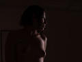 MaryCruzz with dark curly hair, small bare tits, and a tattoo on her left shoulder, standing in dim light, profile view, smiling softly.